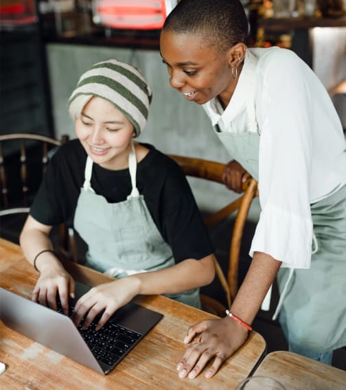 Two women in aprons working on a laptop in a cafe.